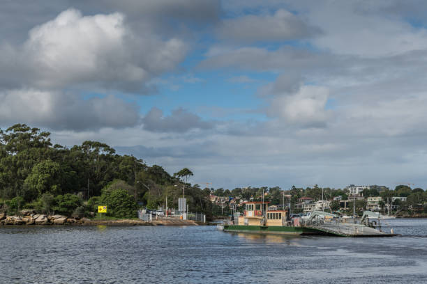 Parramatta, Australia - March 24, 2017: Green-yellow Mortlake ferry crosses Parramatta River transporting cars and people. Houses and green vegetation on shore under heavy cloudscape.