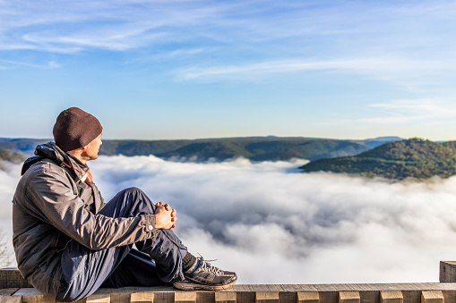 Side profile of man sitting on railing by mountains and floating fog clouds in morning in Grandview Overlook, West Virginia