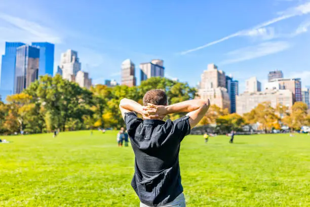 Back closeup of one happy young man with hands behind head, standing in Central Park in New York City during sunny autumn day with skyscrapers buildings and people