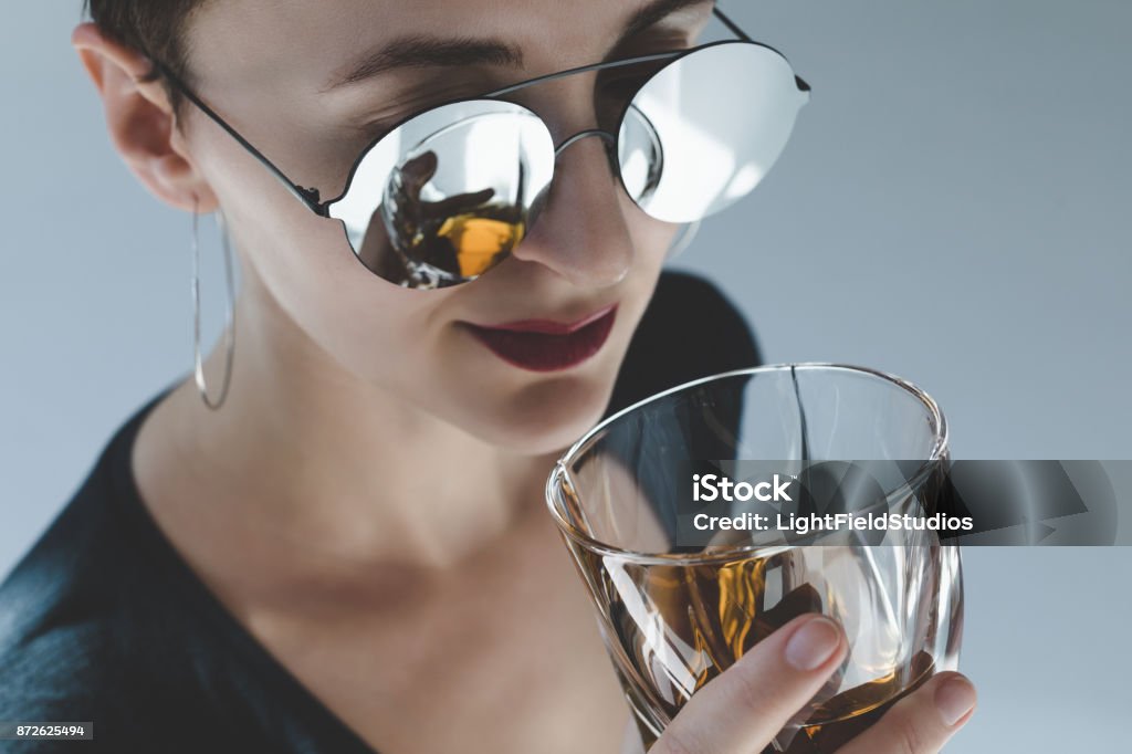 woman drinking whiskey close-up view of beautiful young woman in sunglasses drinking whiskey isolated on grey Whiskey Stock Photo