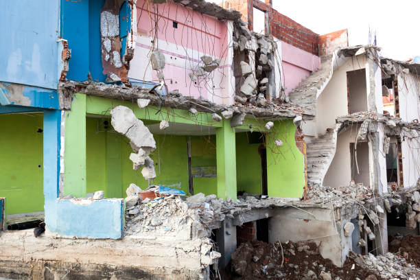 A heap of bricks, wood, and shattered windows marks the remains of a demolished building, with debris spread across bare earth as nearby homes and trees stand untouched in the distance