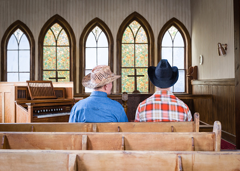 horizontal image of two caucasian cowboys sitting in an old romantic church pew in front of a wall of arched windows and an old organ sitting beside them.