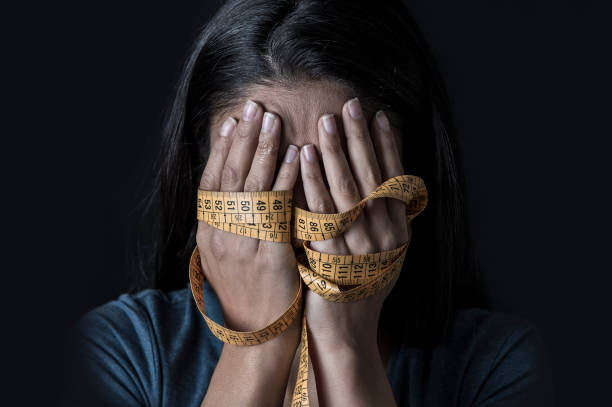close up hands wrapped in tailor measure tape covering face of young depressed and worried girl suffering anorexia or bulimia nutrition disorder on black background stock photo