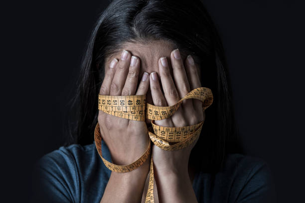 close up hands wrapped in tailor measure tape covering face of young depressed and worried girl suffering anorexia or bulimia nutrition disorder on black background close up hands wrapped in tailor measure tape covering face of young depressed and worried girl suffering anorexia or bulimia nutrition disorder on black background obsessed with diet and overweight body conscious stock pictures, royalty-free photos & images