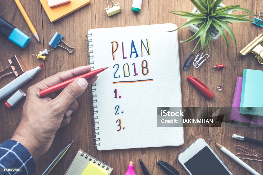 PLAN 2018 message with male hand writing on notepad paper PLAN 2018 message with male hand writing on notepad paper on wooden table and office supplies.Business plan concepts.flat lay design 2018 Stock Photo