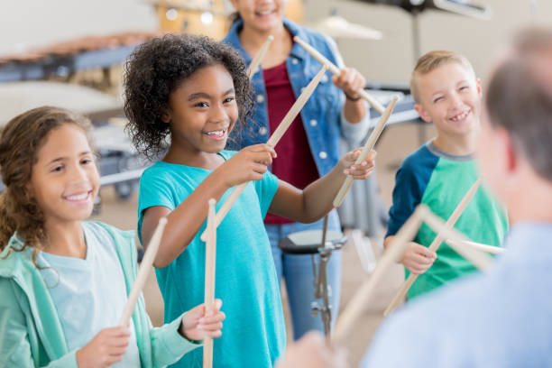 An unrecognizable band teacher stands in front of an attentive group of elementary age children and teaches drumming.  The smiling children look at him and each hold a pair of drumsticks up in the air.
