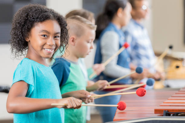 Adorable little girl smiles for camera while playing xylophone In this side view, a cheerful little girl turns to smile for the camera as she plays the xylophone with a little boy.   In the background, other people play a different xylophone. marimba stock pictures, royalty-free photos & images