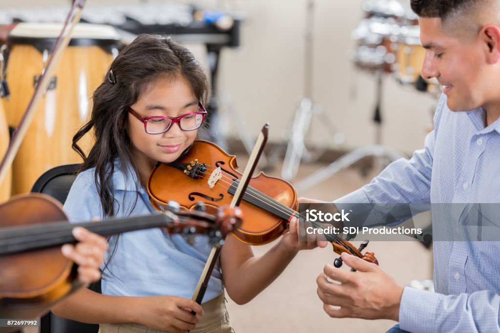 Little girl receives instruction during violin class A smiling little girl sits next to her attentive music teacher and plays the violin during music class.  Her teacher adjusts the violin's position as she plays. Music Stock Photo