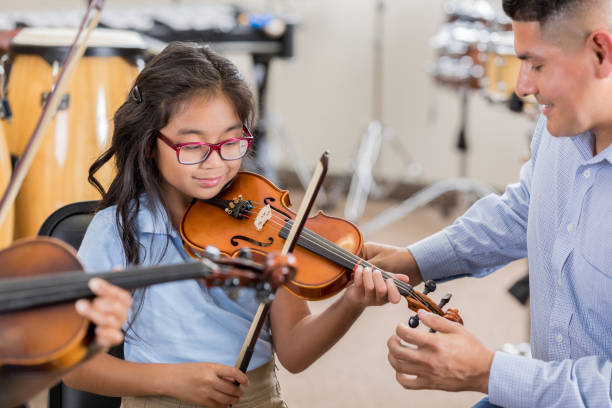 Little girl receives instruction during violin class A smiling little girl sits next to her attentive music teacher and plays the violin during music class.  Her teacher adjusts the violin's position as she plays. music theory stock pictures, royalty-free photos & images