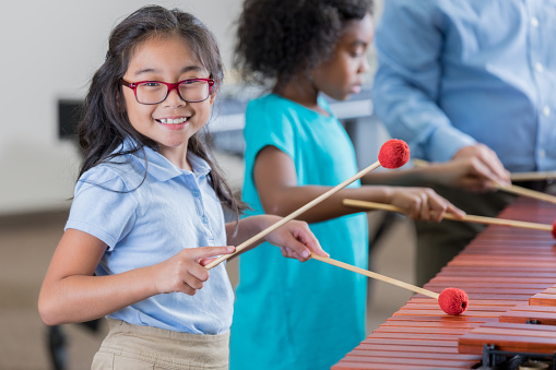 An adorable elementary age girl turns to smile for the camera as she stands beside her unrecognizable teacher and another little girl at a xylophone.  She is learning to play the instrument in band class at school.