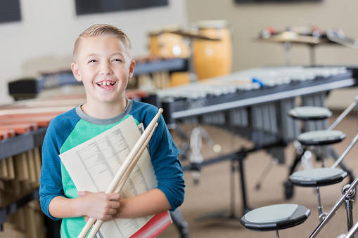 An adorable little boy smiles for the camera as he stands proudly in his music room at school.  He is holding sheet music and a set of drumsticks.