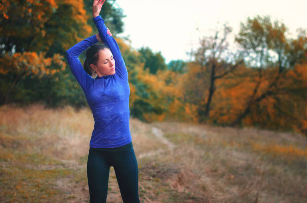 istock caucasian athletic girl in blue shirt  performs warming-up raised her hands up  before jogging on the autumnal fores 872802474
