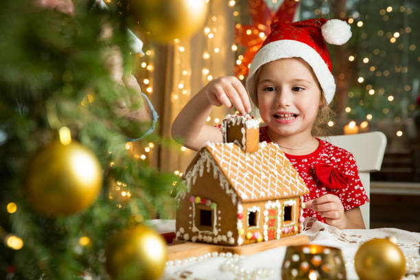 Adorable little girl in red hat decorating Christmas gingerbread house with glaze. stock photo