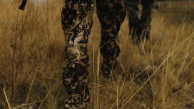 Two bow hunters walk through grass carrying a compound bow, they stop at the top of a large ravine to overlook their hunting ground. The shallow depth of field and slow motion give the shot a feeling of intensity as he walks in full camouflage.