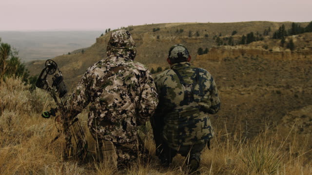 Two hunters sit, silhouetted against the rugged mountain terrain they are hunting. Their camouflage makes they almost disappear into the surroundings.