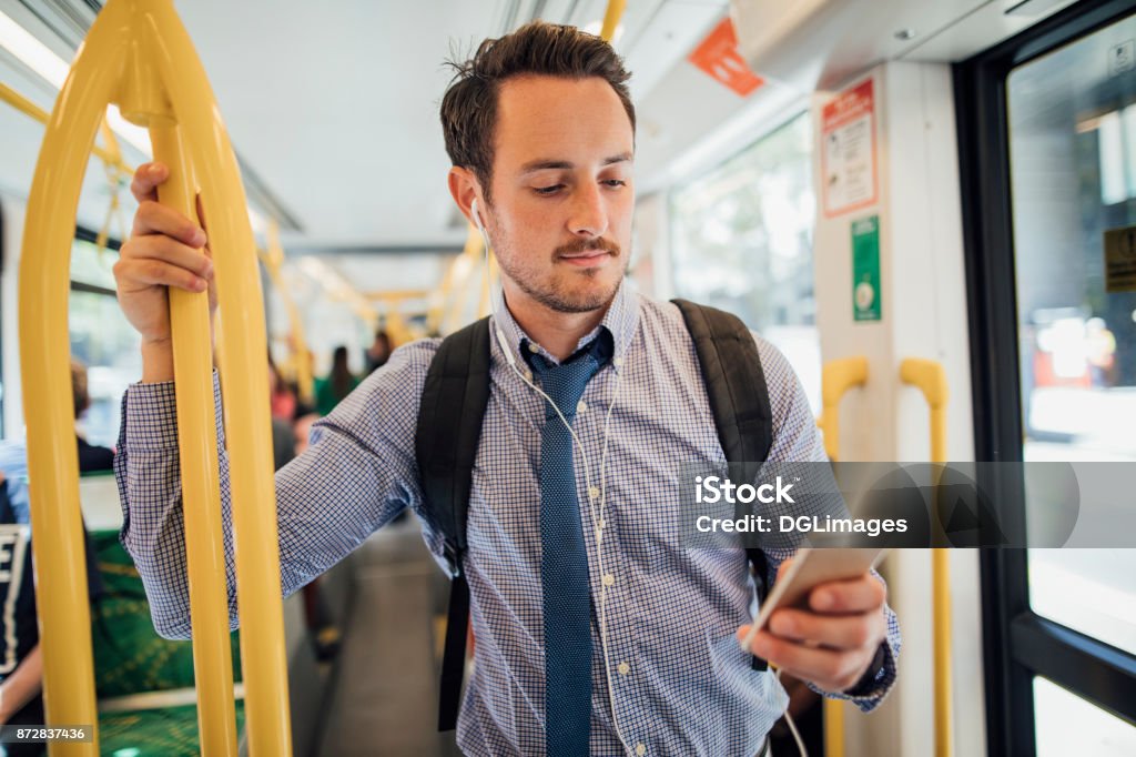 Businessman Commuting By Tram In Melbourne Millennial businessman is commuting on a tram in Melbourne, Victoria. He is watching something on his smart phone with headphones while standing and holding on to the rail. Bus Stock Photo