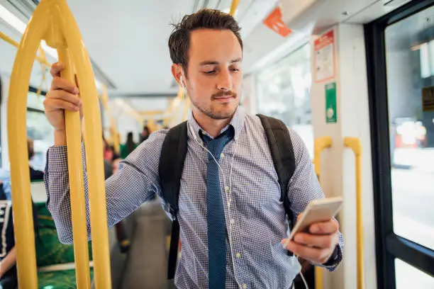 Millennial businessman is commuting on a tram in Melbourne, Victoria. He is watching something on his smart phone with headphones while standing and holding on to the rail.