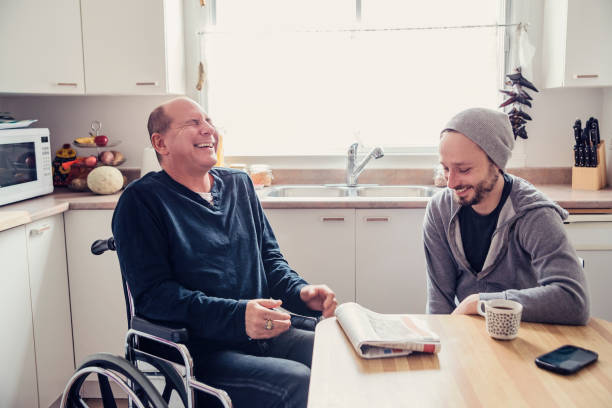 Man visiting a temporary disabled friend and having a coffee. When a friend is hurt and in a temporary wheelchair, it is time to visit and change is mind. Two man having a chat and a coffee in a home kitchen, talking about the morning paper. Horizontal waist up indoors shot in natural light. This was shot in Montreal, Canada. disabled adult stock pictures, royalty-free photos & images