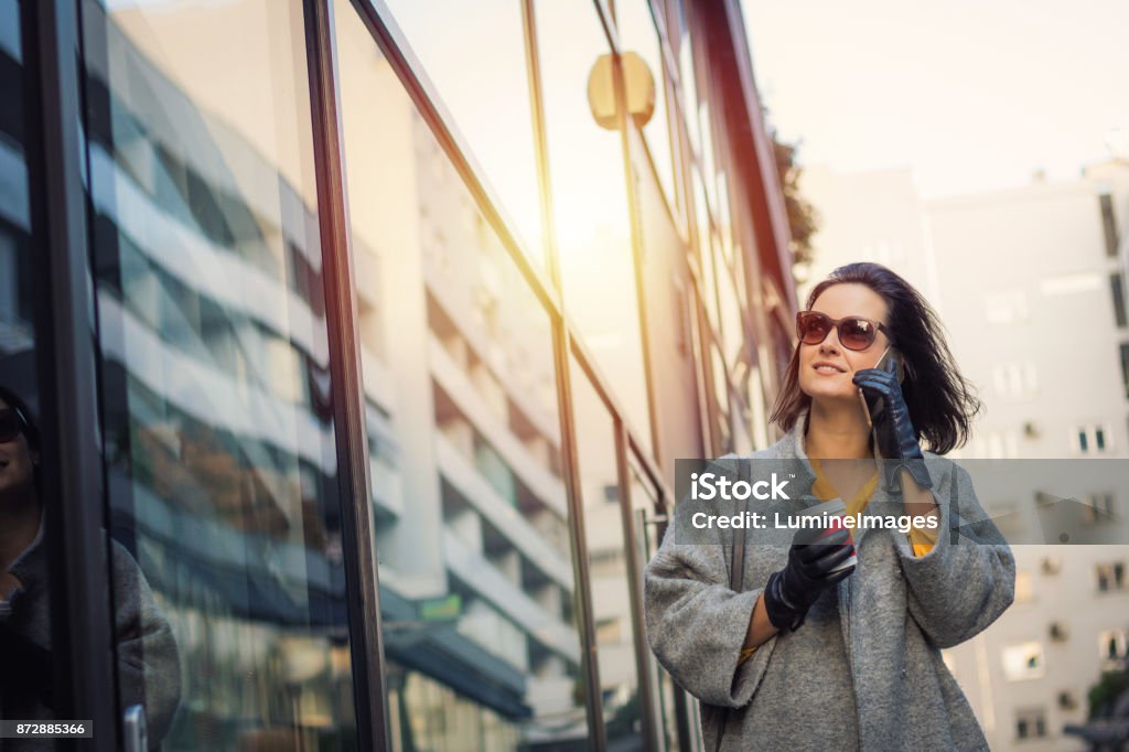 Fashionable woman talking on the phone while window shopping in the city. Beautiful woman walking in the city and talking on mobile phone. Smiling businesswoman with coffee cup walking on the street and making a phone call. Adult Stock Photo
