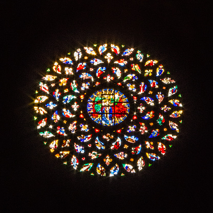 A view from the interior of the church shows a back illuminated round stained glass window with center medallion and sunburst patterned smaller pieces.