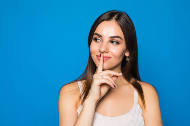 istock Pretty charming young woman having secret while holding finger on lips and showing silence sign while standing on blue background 872926404