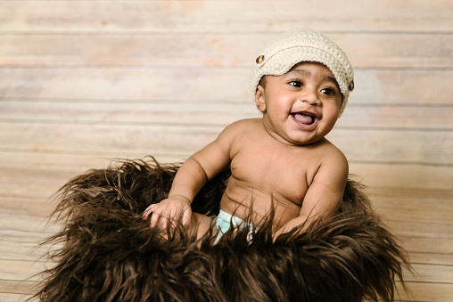 Infant mixed race healthy looking baby boy wearing knitted hat sitting in a fluffy furry basket wooden background modern studio shoot vintage look smiling happy six months old having fun