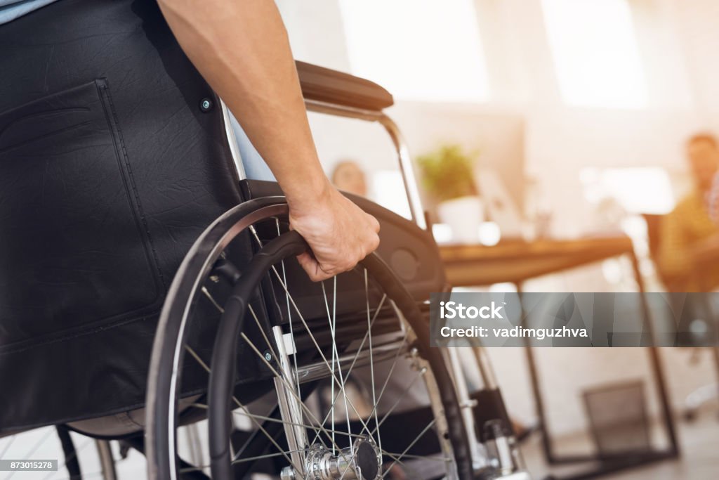 A disabled man is sitting in a wheelchair. A disabled man is sitting in a wheelchair. He holds his hands on the wheel. Nearby are his colleagues Wheelchair Stock Photo