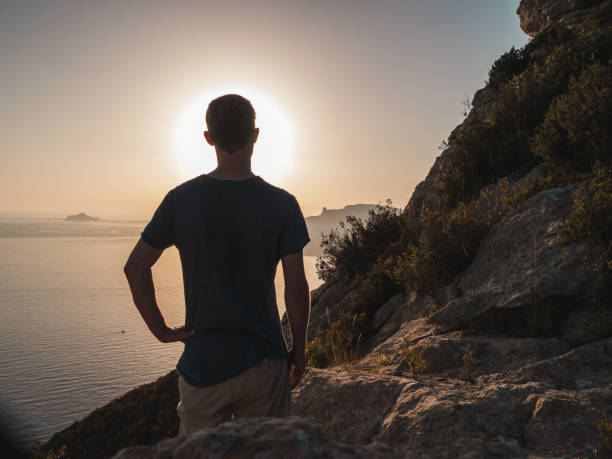 istock Young Man Standing on top of a Cliff, looking at the ocean 873016236