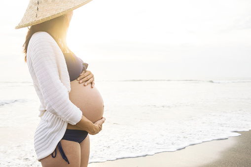 An 8-month old pregnant Asian lady wearing a traditional farmer’s straw hat, contemplates the sunset on a warm tropical beach and holds her belly with two hands. With copy space on the right.