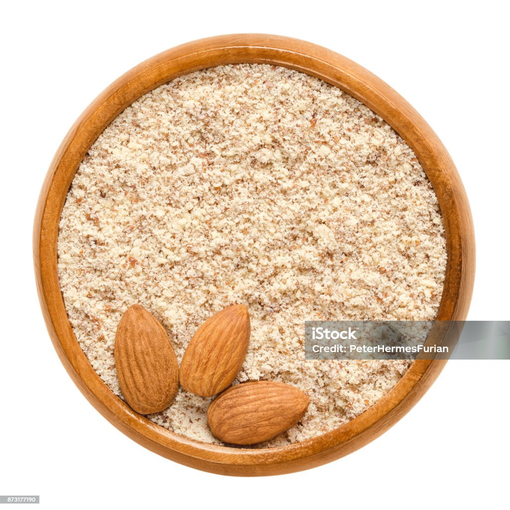 Shelled and ground almond nuts in wooden bowl over white Shelled and ground almond nuts in wooden bowl. Edible, dried, brown seeds of Prunus dulcis. Ingredient in marzipan, nougat, cookies. Isolated macro food photo close up from above on white background. Almond Stock Photo