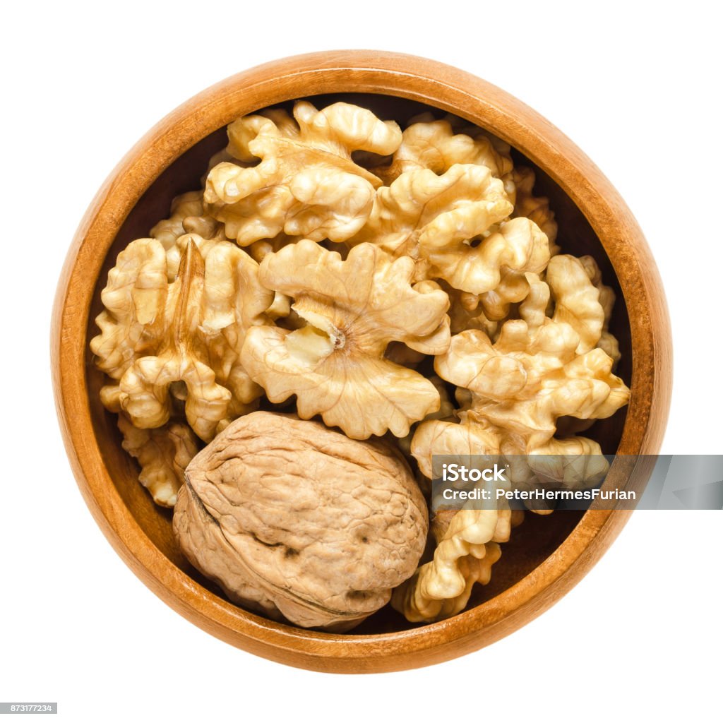 Walnut and shelled walnut kernel halves in wooden bowl Whole walnut and shelled walnut kernel halves in wooden bowl. Seeds of the common walnut tree Juglans regia. Snack or used in bakery. Isolated macro food photo close up from above on white background. Walnut Tree Stock Photo