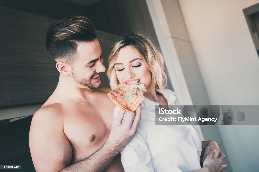 Boyfriend feeding girlfriend with a slice of pizza while hugging her from behind An attractive young boyfriend is seen standing in the kitchen, feeding his girlfriend with a slice of pizza while hugging her from behind. Adult Stock Photo