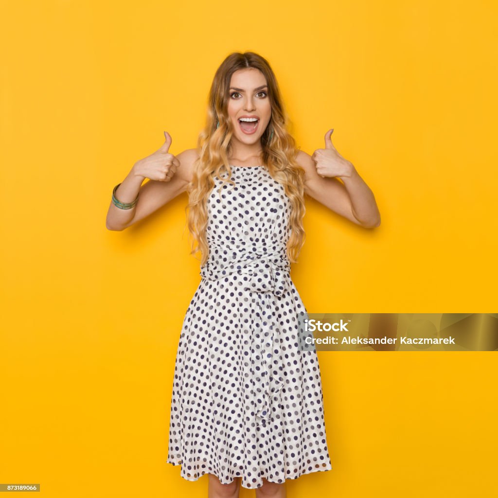 Excited Woman Is Shouting And Showing Thumbs Up Beautiful young woman in white dotted summer dress is showing thumbs up, looking at camera and shouting. Three quarter length studio shot on yellow background. Thumbs Up Stock Photo