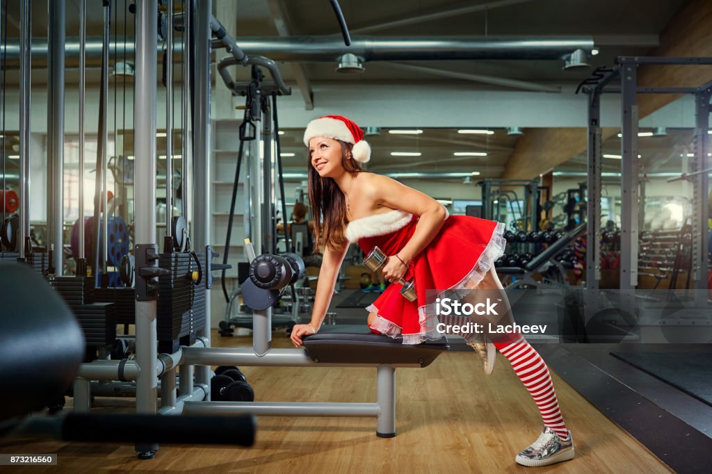 Girl in Santa costume with dumbbells in the gym on Christmas. Girl in Santa costume doing exercises with dumbbells in the gym on Christmas Day. Christmas Stock Photo