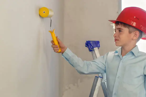 Boy in a red construction helmet paints a wall. Repair in the apartment