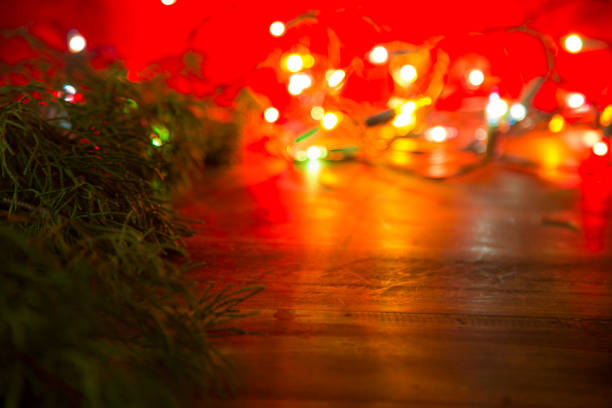 Red and green Christmas holiday background including a green cedar tree branch and white lights on wooden table.  Copyspace at side.