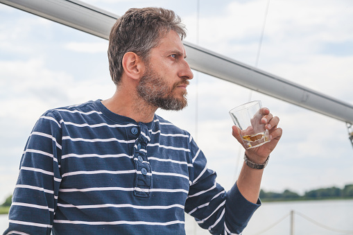 Businessman with a beard in a striped sweater drinks whiskey from a glass on a yacht close-up