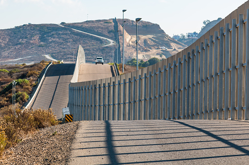 The international border wall between San Diego, California and Tijuana, Mexico, with an approaching U.S. Border Patrol vehicle on a nearby hill.