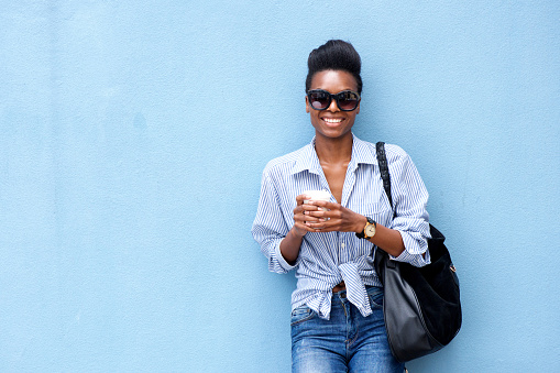 Portrait of smiling young woman leaning against blue wall with coffee cup and purse