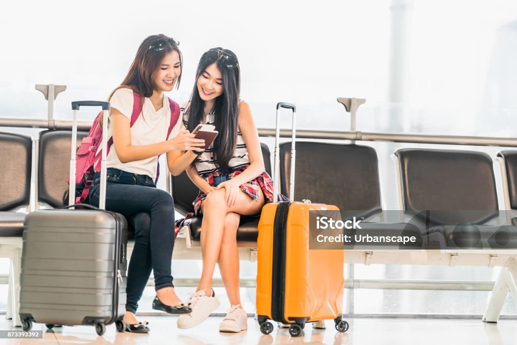 Dois jovens menina asiática usando smartphone verificar voo ou web check-in, sentar no assento de espera do aeroporto. Viagens aéreas, estilo de vida, emocionante viagem de férias de verão ou conceito de aplicativo de dispositivo de telefone móvel - Foto de stock de Aeroporto royalty-free