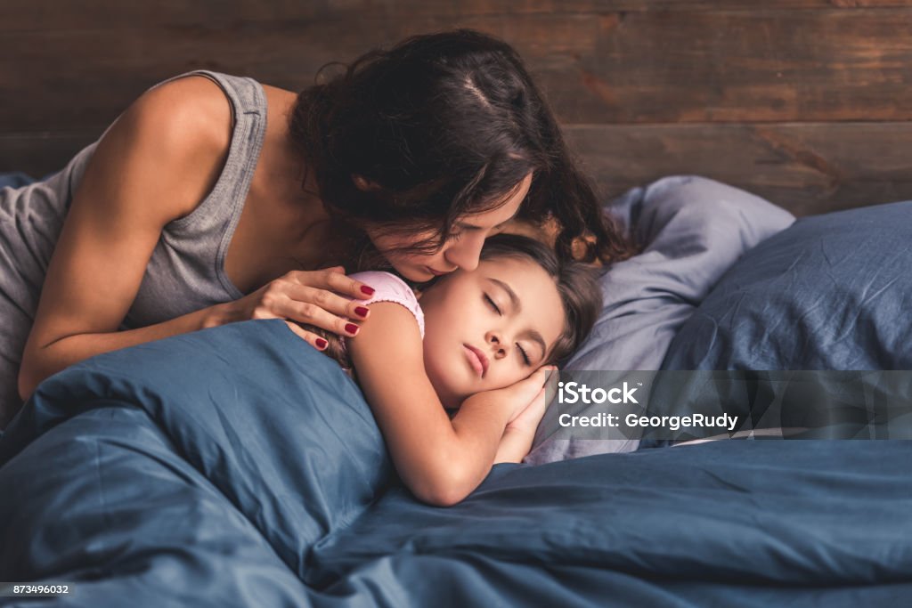 Mom and daughter at home Pretty little girl is sleeping in bed at home, mom is kissing her in cheek Sleeping Stock Photo