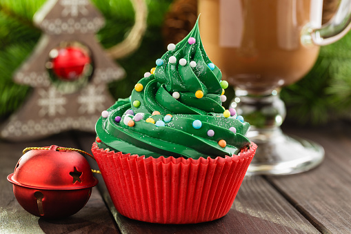 Christmas green cupcake with colorful sprinkles in red cup on wooden background with festive decorations and cup of cocoa