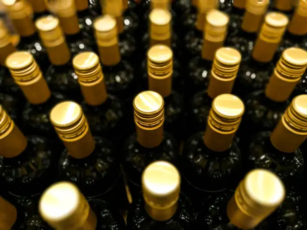 Close up color image depicting an overhead view of a large collection of dark colored glass wine bottles with gold-colored screw caps. Selective focus is on the row of bottles in the middle, while the other bottles are nicely thrown out of focus. Room for copy space.