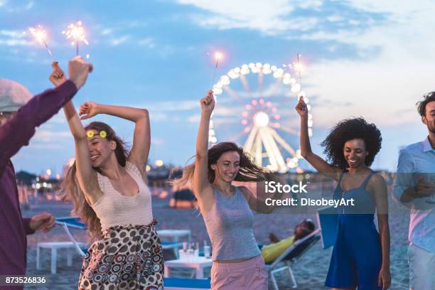 Foto de Felizes Amigos Fazendo A Festa Na Praia Depois Do Anoitecer Com Fogos De Artifício E Bebendo Champanhe Jovens Se Divertindo Dançando Ao Ar Livre Soft Concentrar No Rosto De Garota Centro Juventude E Conceito De Festival e mais fotos de stock de Miami