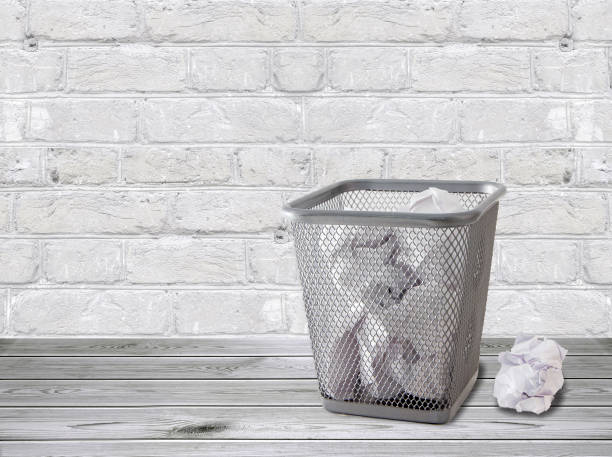 A trash can with crumpled papers stands on the floor against a white brick wall