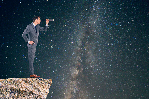 Businessman searching through telescope on rocky cliff against star field background