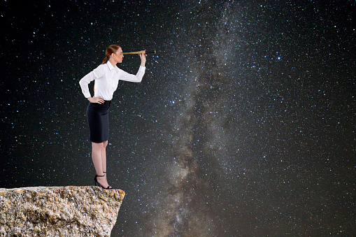 Businesswoman searching through telescope on rocky cliff against star field background