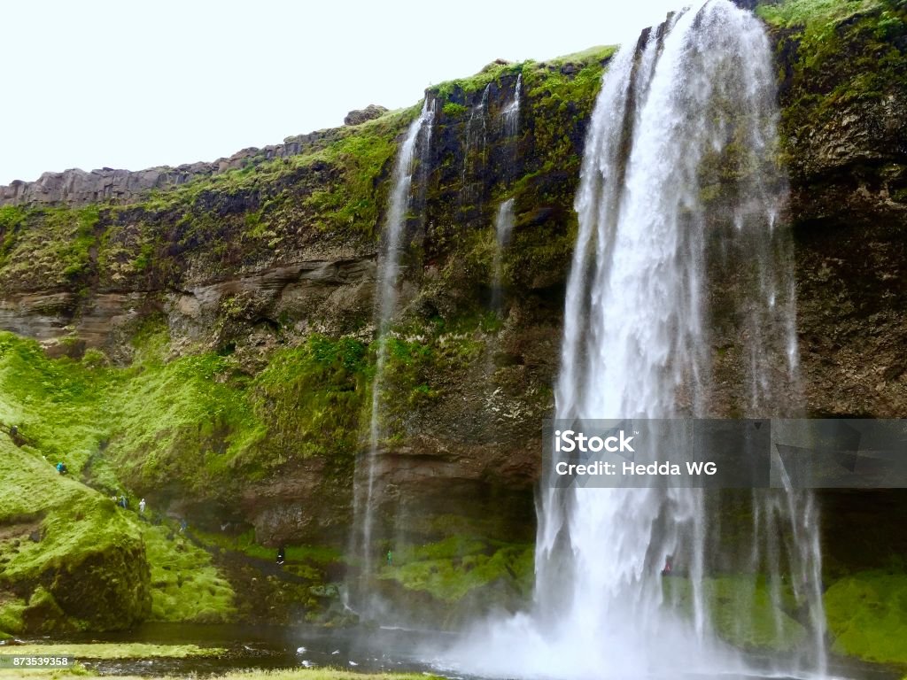 Seljalandsfoss waterfall in Iceland with people walking on a path behind the waterfall Picture of Seljalandsfoss waterfall in Iceland from the side. There is a path behind the waterfall, and people are walking on the path Beauty In Nature Stock Photo