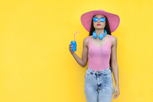 Woman wearing pink vest and colorful sunglasses, blue headphones and holding blue can in front of yellow wall background.