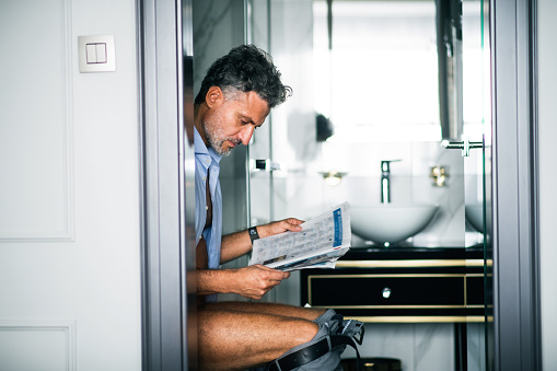 Mature businessman in a hotel room bathroom. Handsome man sitting on the toilet, reading newspapers.
